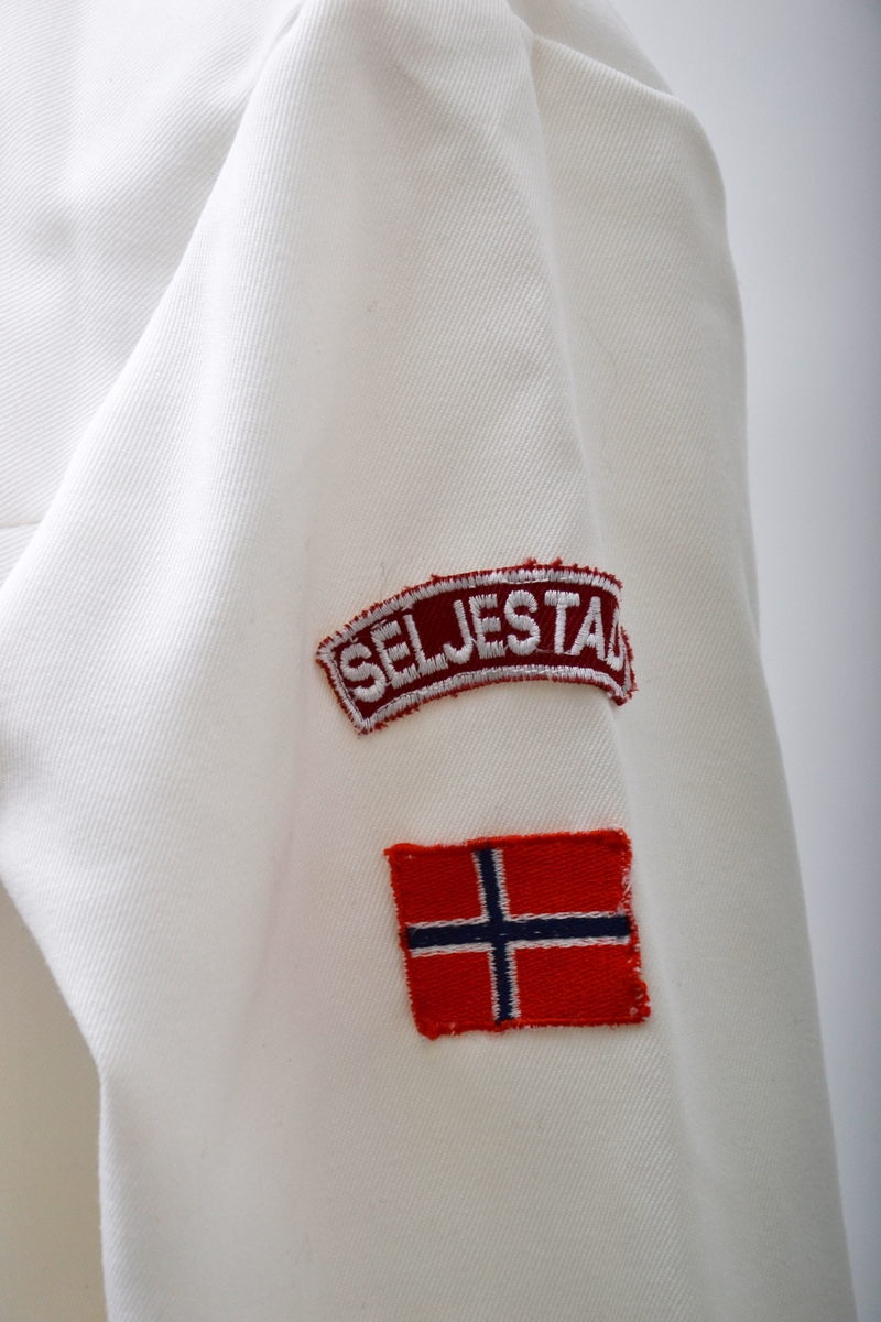 Kort hvit uniformskjole brukt av tambumajor (?) med glidelås i ryggen. Kragen er kantet med rødt bånd. To røde snorer  i sløyfeform er syd fast på brystet. På venstre arm er det sydd fast et norsk flagg og en merkelapp med tekst: "Seljestad". To skulderklaffer med metallknapper.