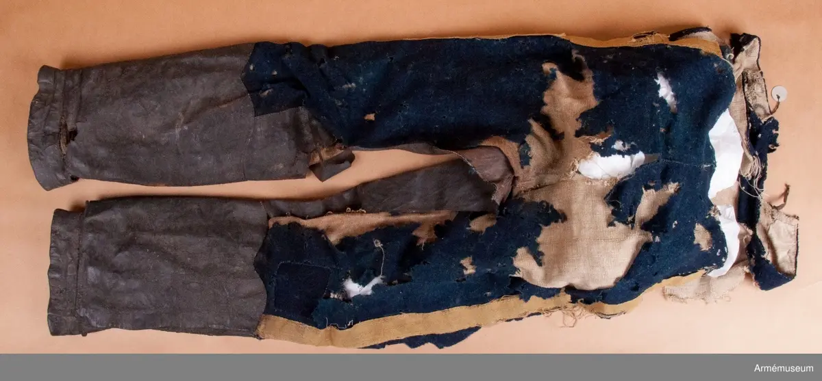 Blå ridbyxor med gula revärer och skinnskodda ben och byxbak.

Byxorna är sydda av ett ganska tunt blått kommisskläde, och är helfodrade med oblekt linne- eller hampalärft.
Modellen är konstruerad med två fram- och två bakstycken, och har hel lucka fram som har knäppts mot en gördel eller linning. Luckan har 5 knapphål fram, varav tre har knäppts mot gördeln. I luckans sidor upptill sitter två spänntampar för knäppning bak i ryggslutet. I sömmen bak sitter en infälld kil för reglering av midjevidden, samt en hank av läder för upphängning.
I sidan har byxorna revärar av 4 cm breda gula redgarnsband.
Byxbenen är skodda med brunt skinn i en höjd av ca 36 cm nertill. Kanten är formad i två bågar fram och en spets bak. Skinnskoningen fortsätter på baksidan upp på insidan av låren och i byxbaken.
Byxorna är mycket trasiga och malätna, och både yttertyg och foder har lagats ett flertal gånger under deras användningstid.
Samtliga knappar saknas förutom i höger byxben längst ner där det sitter rester av enkla skinnknappar i varje sida.

Märkt på insidan "LS No 2".