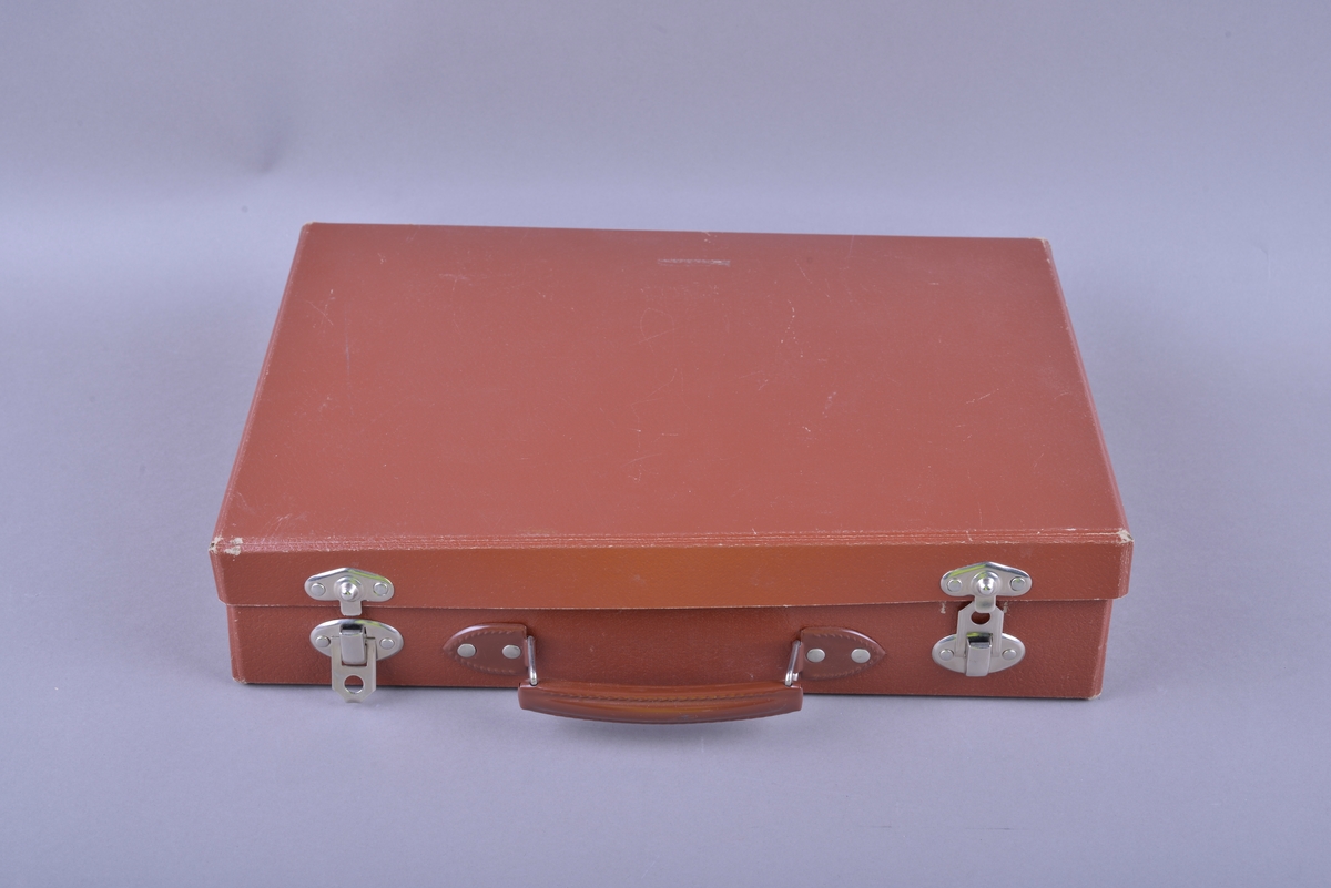 Rektangulær brun koffert av papp, med håndtak og lås. Inne i kofferten er det små pappesker. Pappeskene inneholder små treklosser, plater med pålimte miniatyr-leker. Blant annet dukke, hund seng, saks, pipe. I tillegg er det flere små knapper, notatblokk og tekstilbånd. I kofferten ligger også en bok merket "TERMAN-MERRILL // NORSK STANDARD".