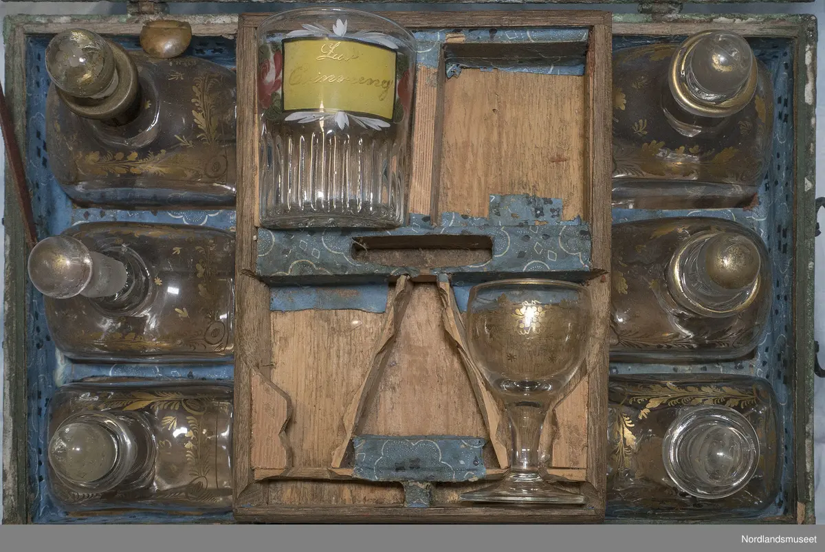 Velbevart flaskefor. Rektangulært skrin med flat lokk med rom for seks store flasker på sidene og seks små flasker (flakoner) i midten. Samtlige dekorert med floralt dekor i gyllen farge. Over småflaskene et brett med utforminger til opprinnelig to begerglass og tre små stettglass. Bare ett glass av hver sort bevart. Kassens innside er foret med blått dekorert papir. En rød filt er limt på innsiden av lokket for å polstre glassene.