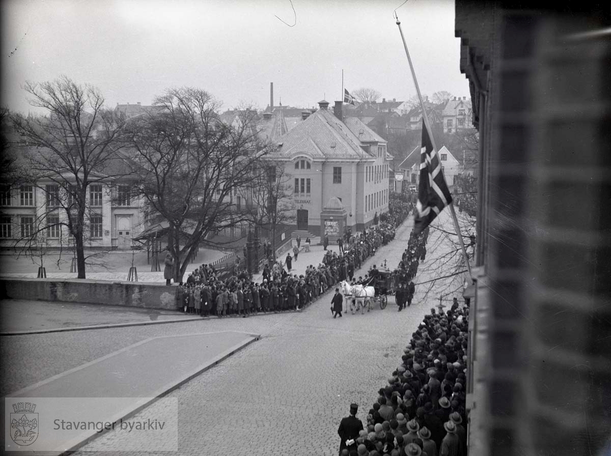 Vognen med kisten leder an begravelsesfølget. Publikum ser på langs posthuset, Kongsgård, Torget og fengselet. ...Lars Oftedal gravlegges.