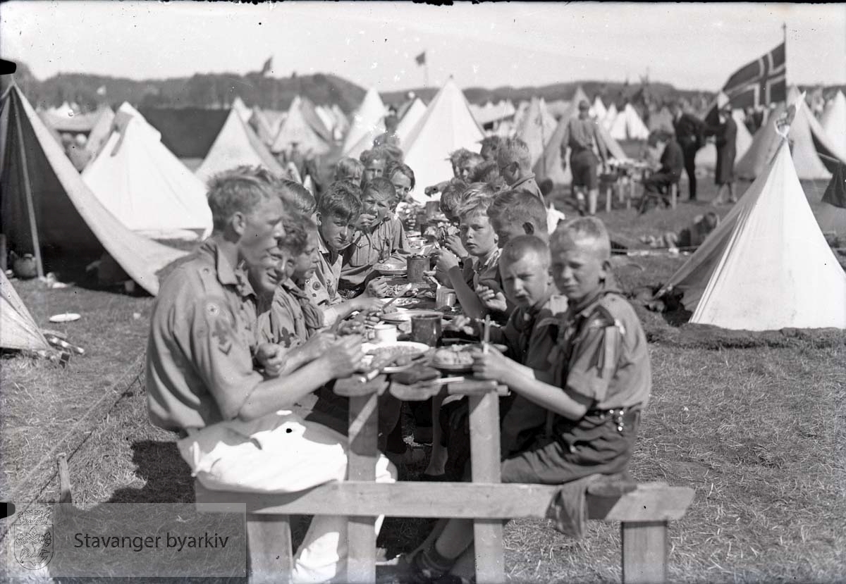 Speidere har måltid i leiren. Telt oppstilt på rekke og rad i bakgrunnen.