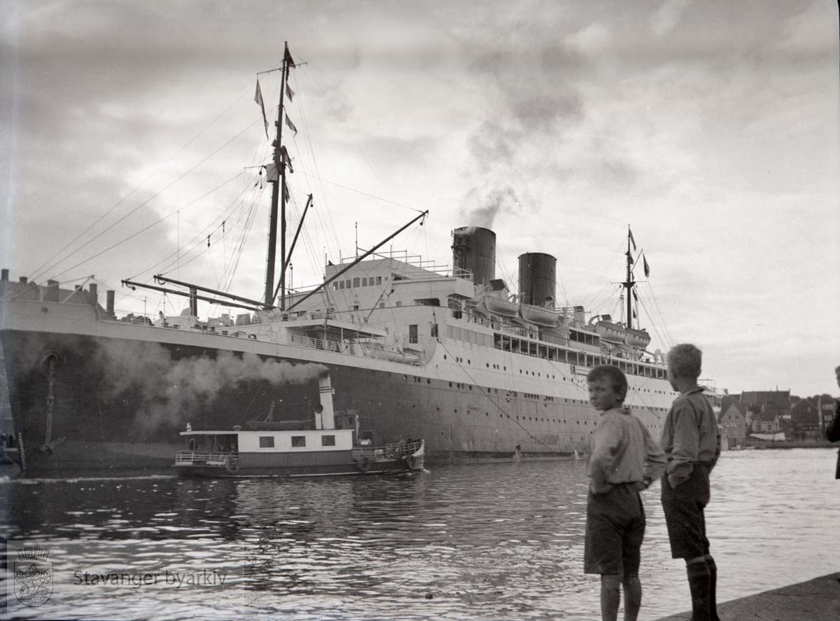 D/S "Colombie" i Vågen..Skipet ble bygget i 1931 for trafikk mellom Frankrike og det karibiske området.. .Eieren var det kjente rederiet Compagnie générale transatlantique (CGT eller "French Line"), som i årene fram til 2. verdenskrig også benyttet "Colombie" til sommercruise i nordiske land.. ."Colombie" besøkte Stavanger flere ganger på 1930-tallet. Skroget ble hvitmalt i 1935. Fotoet er sannsynligvis tatt ved det første anløpet lørdag 19. august 1933. (Se Stavanger Aftenblad for 21. august 1933 med foto av "Colombie" ved Strandkaien; babord anker er låret nesten til vannlinjen - som på arkivfotoet.). .Aftenbladet beskriver et anløp med forviklinger, avlyst tur til Dirdal og "tiggende" barn på kaien ("en skam for byen"). Guttene på arkivfotoet er kanskje blant dem som ble bortvist av politiet?. .Båten foran "Colombie" er buøyfergen "Buøy I" som gikk til Nyhavn...(Opplysninger gitt av Einar Haualand, 05.09.2022).