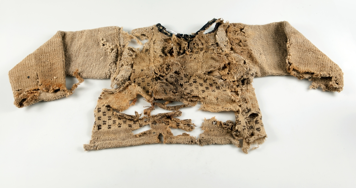 Fragment av stickad tröja med mönsterstickad bård i brunt och beige ullgarn. Bålen i 1-trådigt ullgarn mönsterstickad i rutmönster i brunt, natursvart och beige. Ärmarna är rundstickade i 2-trådigt ullgarn i naturvit, natursvart, brunt och beige. Sprund mitt fram 10 cm långt. Halsurringning och sprund mitt fram kantad med samma tyg som finns som lagningar på UM23252 och A 38 (UM ?). Hyska och hake i mässing mitt fram.