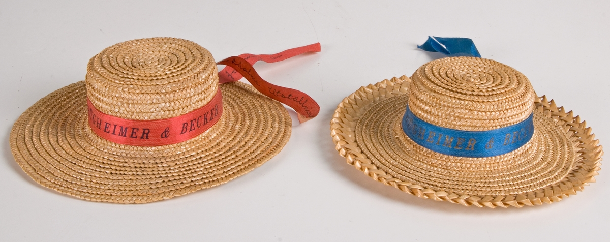 Två dockhalmhattar med rött respektive blått band.
a) Med flätat kant och blått band med med guldtexten: "RÜGHEIMER & BECKER, 1897, Minne af Stockholms utställning." Diam 14,5cm.
b) Med rött band och svart text: som a. Diam 14,5.