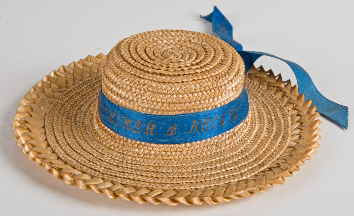Två dockhalmhattar med rött respektive blått band.
a) Med flätat kant och blått band med med guldtexten: "RÜGHEIMER & BECKER, 1897, Minne af Stockholms utställning." Diam 14,5cm.
b) Med rött band och svart text: som a. Diam 14,5.