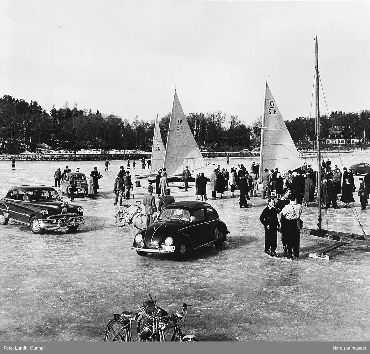 Folksamling på isen på en sjö i Stockholmstrakten år 1954. Människor står krin isjakter, en del personer har skridskor. På isen är bilar parkerade, till vänster en Pontiac årsmodell 1952, till höger en Volkswagen Golf.  Närmast i bild står mopeder parkerade.