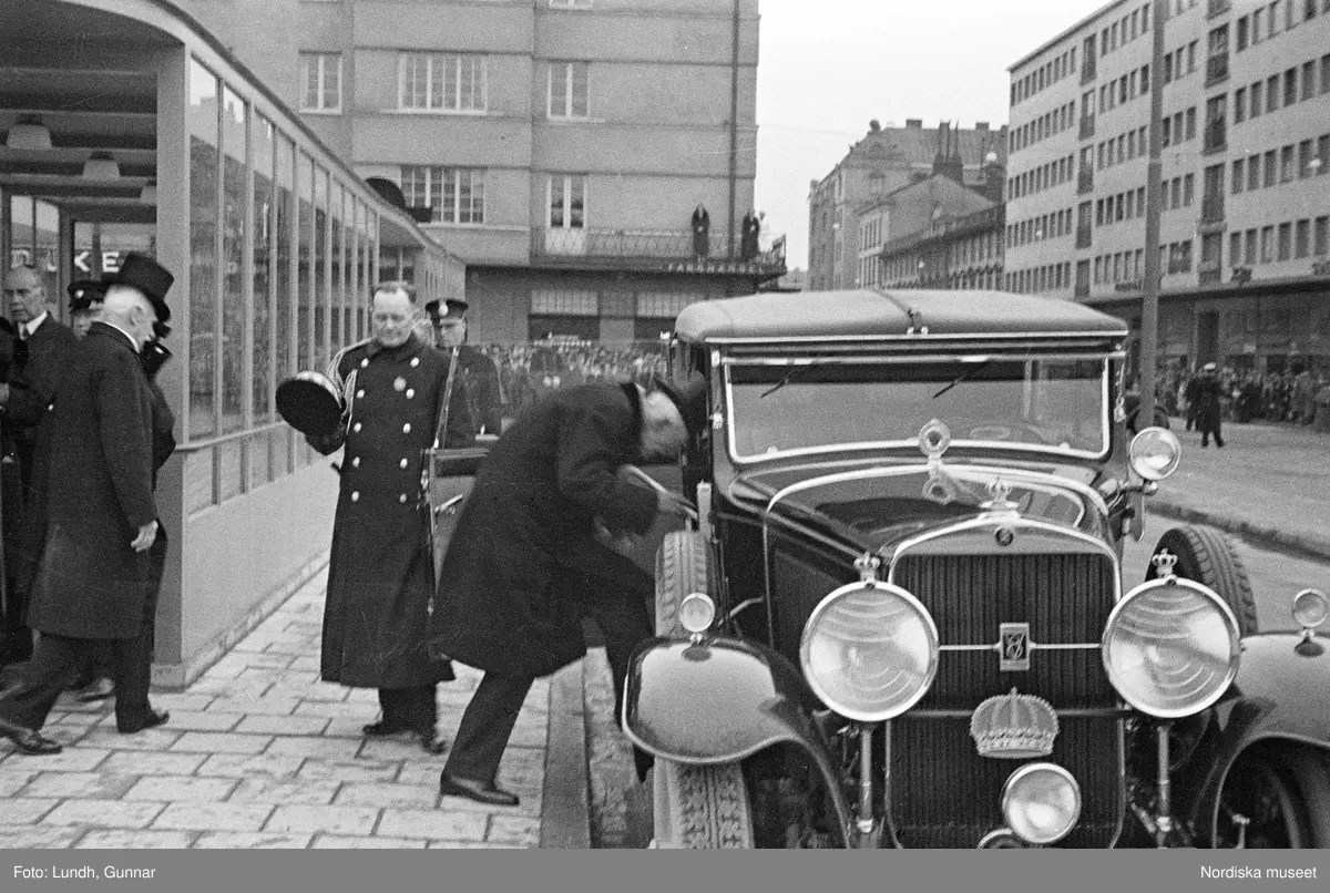 Invigning av Södra Bantorgets (Medborgarplatsen) tunnelbanestation på Södertunneln den 30 september år 1933, Kung Gustaf V kliver in i en bil, porträtt av en man i uniform - en chaufför vid Kungliga Hovstallet, detalj av en bil med lykta och skärm, en grupp män går ut ur en tunnelbanenedgång på Södra Bantorget - män i uniform gör honnör och i bakgrunden står människor i öppna fönster.