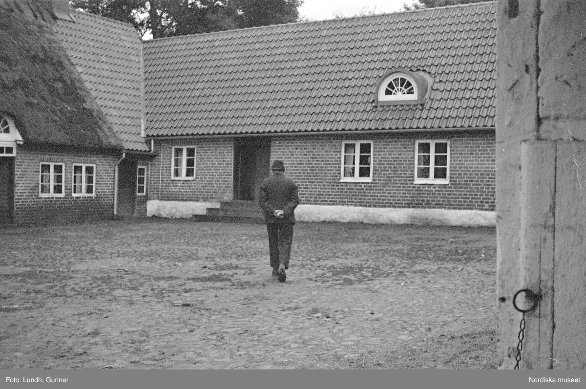 Miljöbilder  och porträtt av Nils och Fredrik Åkesson gåsupfödare i Veberöd, exteriör av bondgård, en hund på en gårdsplan, en flock gäss på en gårdsplan, porträtt av två män och en hund som står på en gårdsplan, en kvinna står i en dörröppning vid en tvådelad dörr, en man står vid en delad ladugårdsdörr, vy över en väg med hagar och stängsel av brädor.