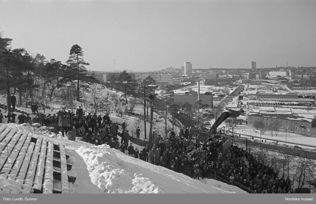 En tävling i backhoppning i Hammabybacken, Stockholm, backhoppare hoppar från en hoppbacke inför åskådare och domare, vy över Södra Hammabyhamnen och Södermalm.