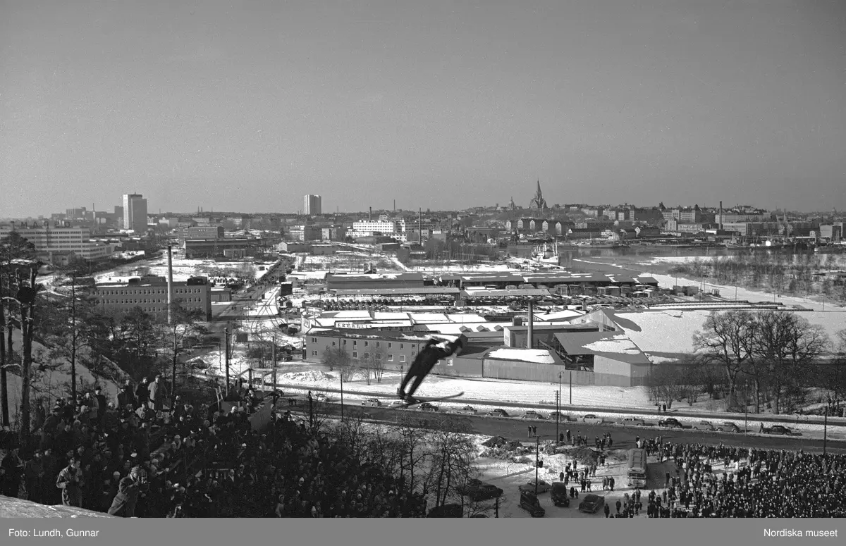 En tävling i backhoppning i Hammabybacken, Stockholm, backhoppare hoppar från en hoppbacke inför åskådare och domare, vy över Södra Hammabyhamnen och Södermalm.