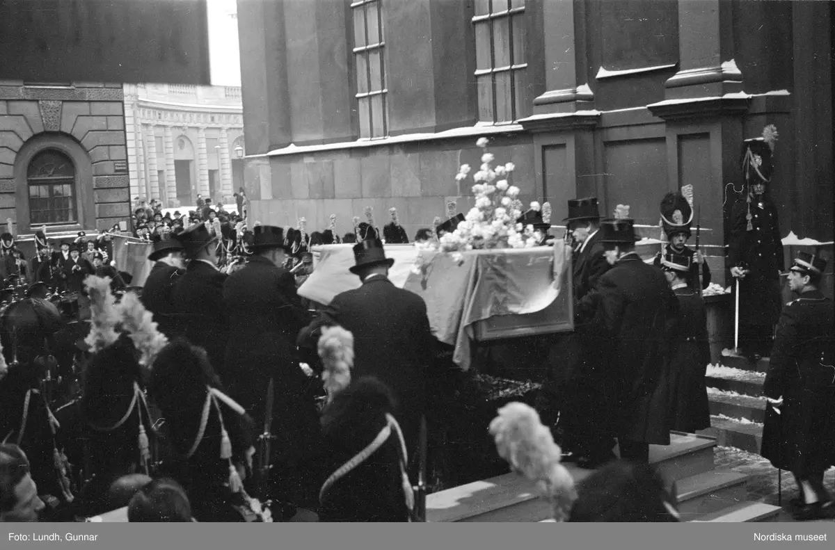 Troligen begravningen av Prins Gustaf Adolf, en kista bärs ut från Storkyrkan, kungafamiljen går ut från Storkyrkan, militärer i uniform står i givakt, kistan står på en katafalk som dras av hästar, en folksamling står längs vägen när begravningsföljet passerar.