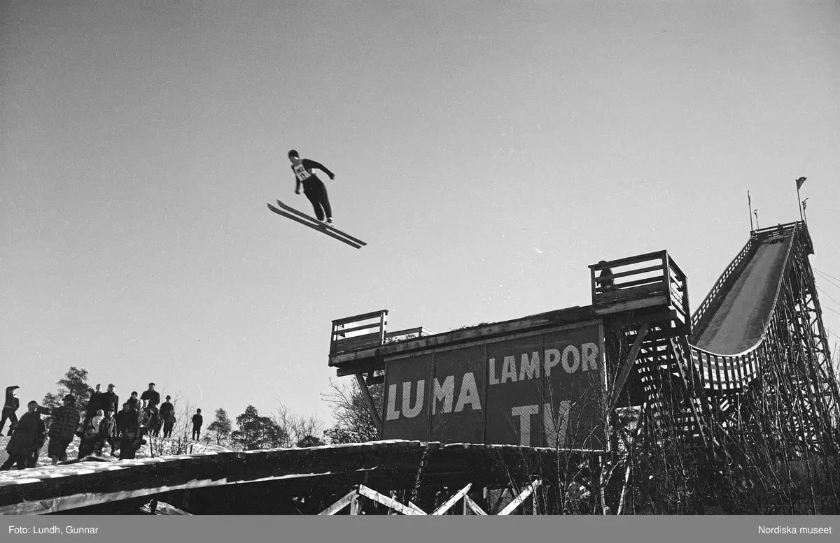En tävling i backhoppning i Hammabybacken, Stockholm, backhoppare hoppar från en hoppbacke inför åskådare och domare, vy över Södra Hammabyhamnen och Södermalm.