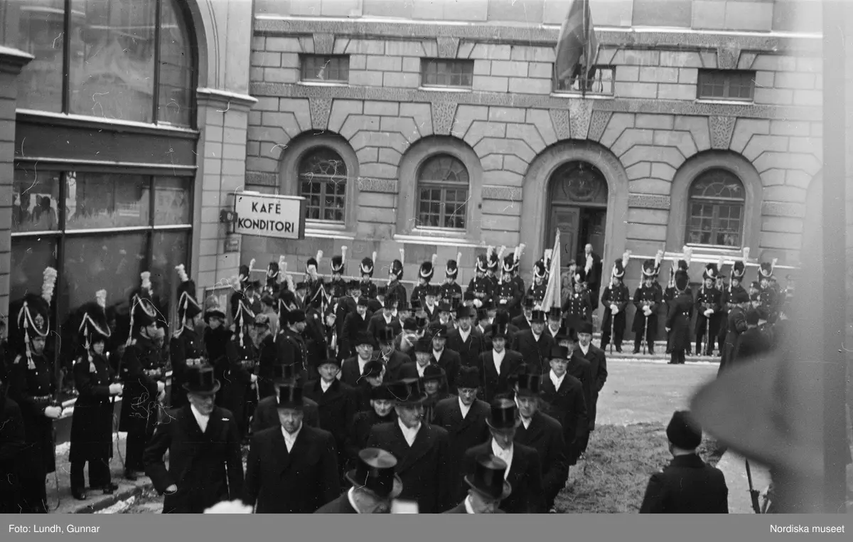 Troligen begravningen av Prins Gustaf Adolf, en kista bärs ut från Storkyrkan, kungafamiljen går ut från Storkyrkan, militärer i uniform står i givakt, kistan står på en katafalk som dras av hästar, en folksamling står längs vägen när begravningsföljet passerar.