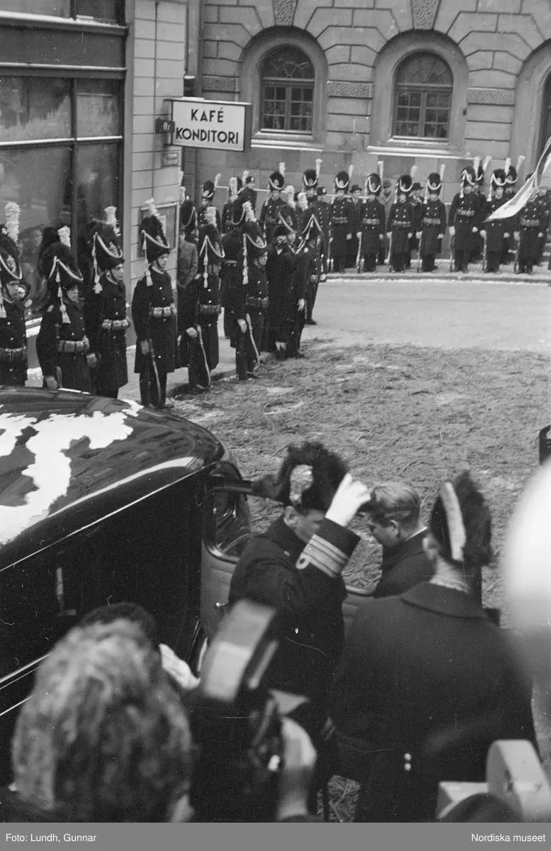 Troligen begravningen av Prins Gustaf Adolf, en kista bärs ut från Storkyrkan, kungafamiljen går ut från Storkyrkan, militärer i uniform står i givakt, kistan står på en katafalk som dras av hästar, en folksamling står längs vägen när begravningsföljet passerar.