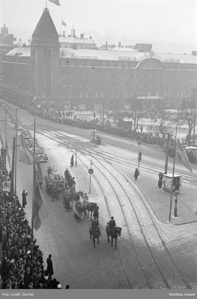 Troligen begravningen av Prins Gustaf Adolf, en kista bärs ut från Storkyrkan, kungafamiljen går ut från Storkyrkan, militärer i uniform står i givakt, kistan står på en katafalk som dras av hästar, en folksamling står längs vägen när begravningsföljet passerar.