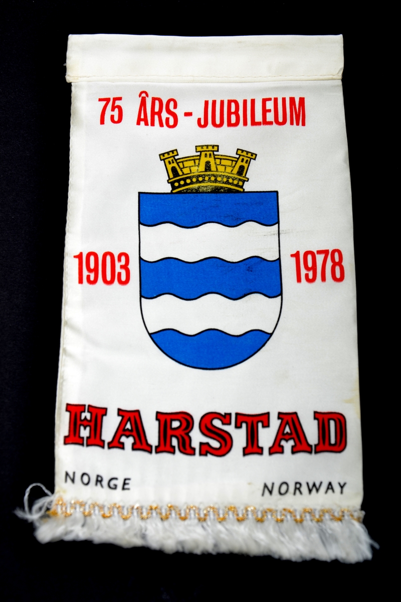 Bordfane fra Harstad kommune tilvirket av hvitt kunststoff med kommunevåpen og en hvit frynsekant nede på vimpelen, uten stativ