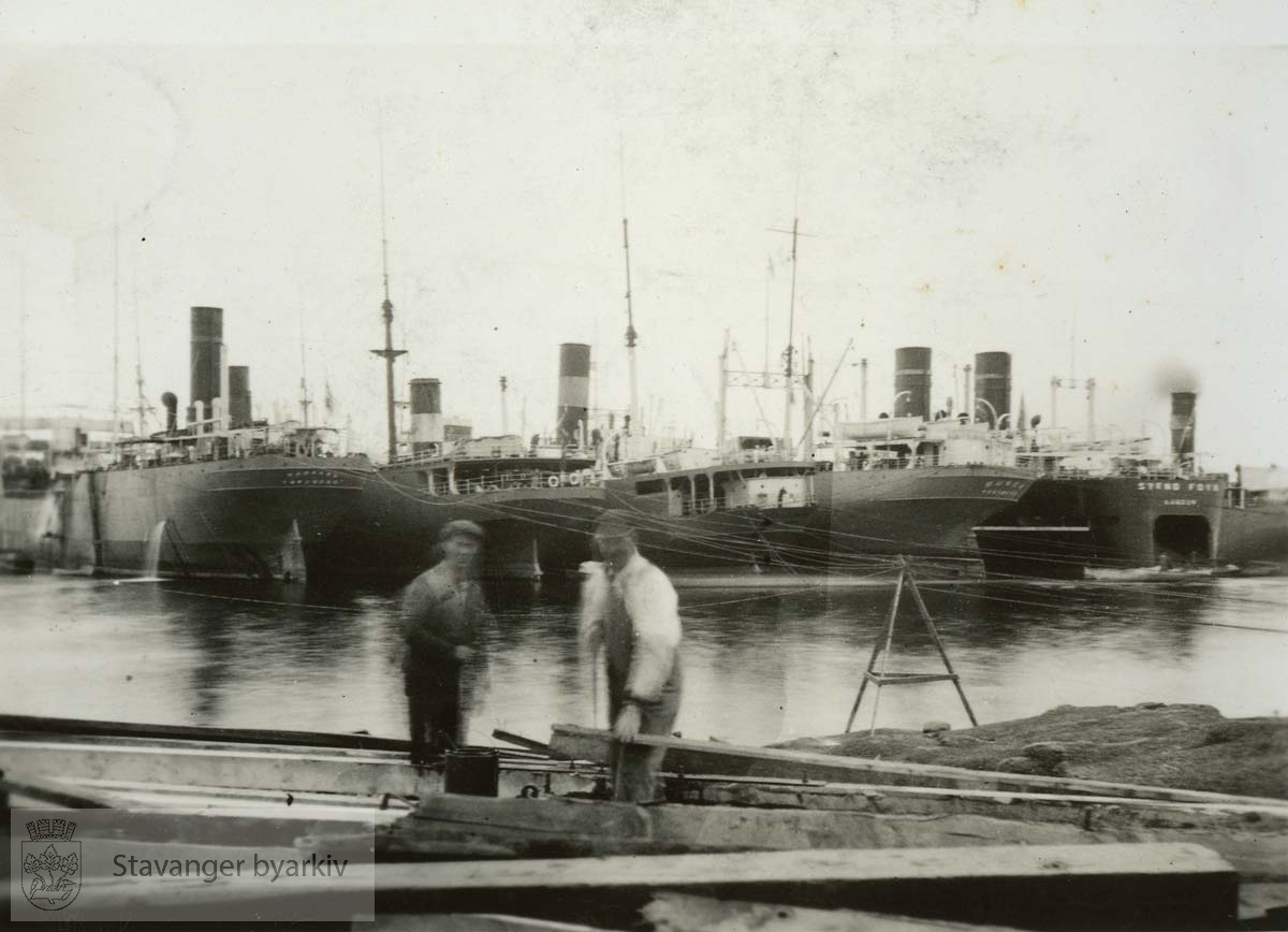 To arbeidere i forgrunnen..Skipet helt til høyre er hvalkokeriet Svend Foyn, bygget i 1931 for Hvalfangerselskapet Sydhavet A/S, men solgt i 1932 til Star Whaling Co Ltd. Det later til at skipet på bildet har hjemhavn London, så med all sannsynlighet er fotografiet tatt etter 1932, muligens i Sandefjord eller Tønsberg