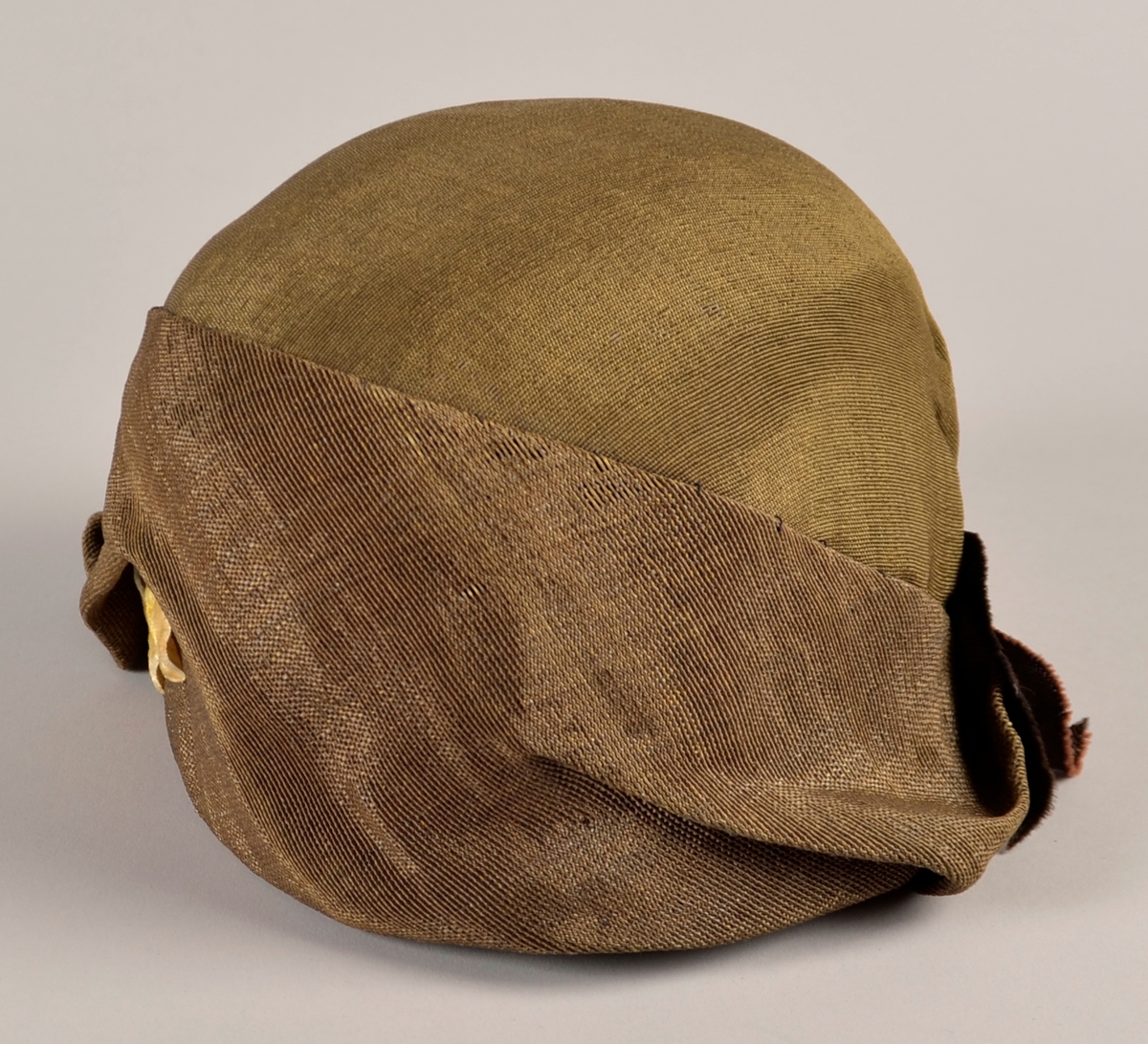 Kvinnehatt. Stiv materiale. Utsida i gyllent, svart/gull, stoff. Sløyfe på sida av brunt band. To gule blad av fløyel til pynt på andre sida. Svart fôr.