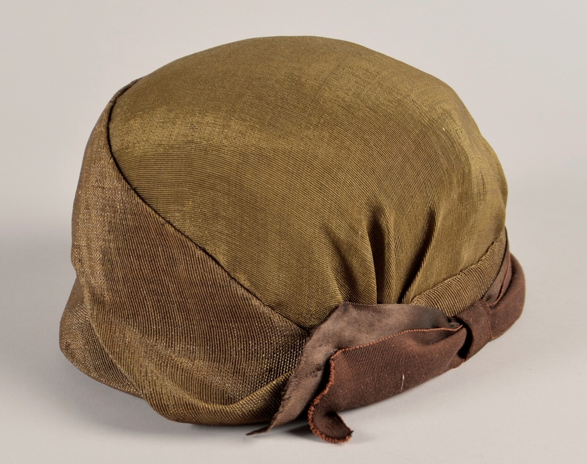 Kvinnehatt. Stiv materiale. Utsida i gyllent, svart/gull, stoff. Sløyfe på sida av brunt band. To gule blad av fløyel til pynt på andre sida. Svart fôr.