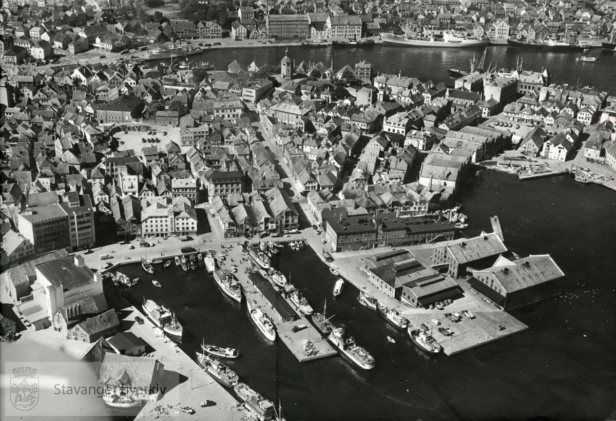 Steinkarkaien, Kjeringholmen med slakthuset, Bredgaten. I bakgrunnen Vågen med Strandkaien og Sandviken...Denne serien med flyfoto fra K. Harstad, har kommunen kjøpt av Johan Ottesens Fotoarkiv. ..Karl Harstad, som grunnla K. Harstad kunstforlag (Oslo Prospektkortfabrikk) i 1925, var den første sivile flyfotoprodusenten i Norge. Som postkortprodusent var Harstad opptatt av nhye produkttyper. I 1929 dannet han sammen med andre selskapet FotoFly og kjøpte fly i Tyskland. Selskapet skulle drive både passasjerflyging og fotograferin. I 1929 fotgraferte selskapet de fleste byene langs kysten av Norge, også i Rogaland. Postkortene med flyfotografi ble produsert med navnet Flyfoto, senere med navnet K. Harstad. En annen stor K. Harstad-postkortserie fra Rogaland skriver seg fra flyfotografering 1950-1951. Da fotograferte firmaet både byer og mindre tettsteder og bygder i fylket. Det er uvisst hvor omfattende K. Harstad sin flyfotoproduksjon var i Rogaland før firmaet ble nedlagt ved overgangen til bruk av fargefoto tidlig på 1960-tallet. Så langt man kan se av postkortsamlinger ved kulturverninstitusjonene og hos private, viser de aller fleste kortene byer og tettsteder, ikke enkelteiendommer og bedrifter.