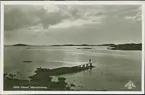 Vykort med motiv från Hönö Hästen. Fotografiet är taget från land ut över en strand och det omkringliggande skärgårdslandskapet. På en brygga ut i vattnet går en person ut mot vattnet.