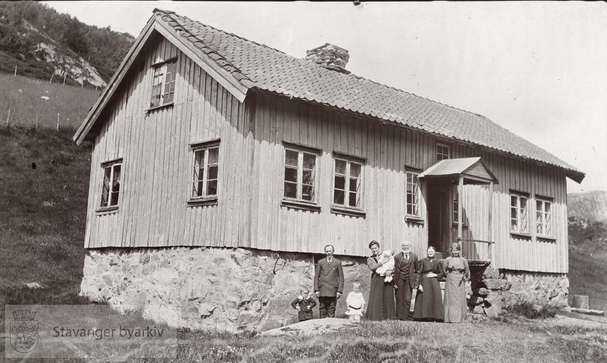 Huset, med tilnavnet "Der heima", var en av tre utskilte gårder etter det som hadde vært én gård, Hammersmark-gården ved Tonstad, med gårdsdrift fra 1630...De voksne er fra venstre gårdbruker og emissær Torkel Hammersmark, og hans kone, Inger Marie Stålesdatter Røinestad, gift Hammersmark. På hennes høyre side står Torkels foreldre, Torje Torjesen Hammersmark og Borgny Mikkelsdatter Espetveit, gift Hammersmark. Ytterst til høyre står Torkels søster og deres datter, Gudrun Hammersmark..Barna er fra venstre Tom og Sverre, og på morens arm, den yngste sønnen Bjarne. .Bildet er tatt på Hammersmark i 1915." ..(Informant: Marit Hammersmark).