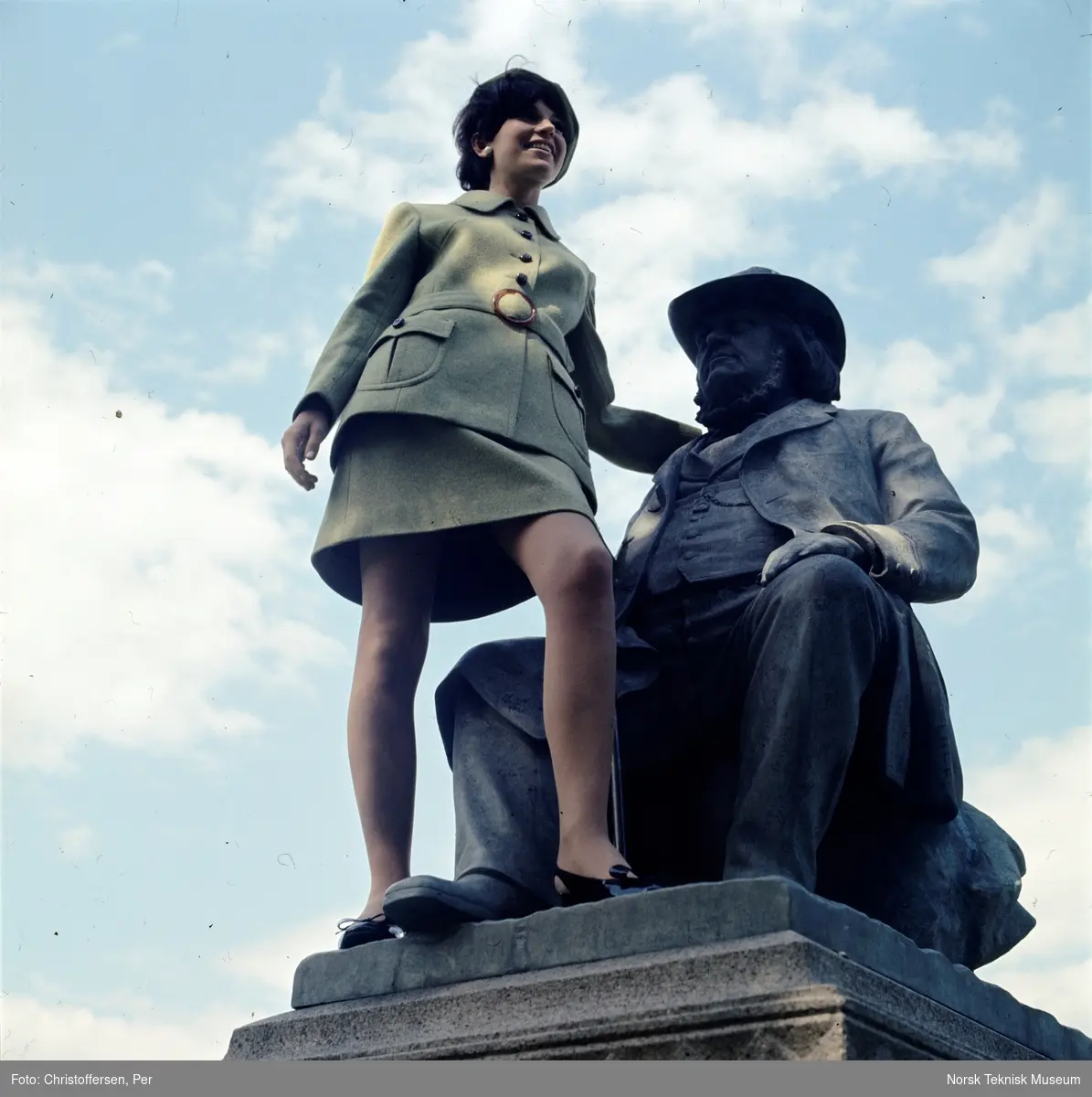 Motebilde, kvinne i grønn drakt, sittende på statuen av eventyrfortelleren Peter Chr. Asbjørnsen (1891) av Brynjulf Bergslien på St. Hanshaugen i Oslo, 1968. Bildet er en del av en serie der noen er publisert i ukebladet Det Nye samme høst med billedteksten: "Øverst til venstre ser du en grønn melert drakt av 70% ull og 30% nylon/rayon. Modellen har svakt skrådd skjørt og lang jakke med belte, utenpålommer med runde hjørner og sesongens tradisjonelle skilpaddespenne og knapper. Modellen koster ca kr. 400,-"