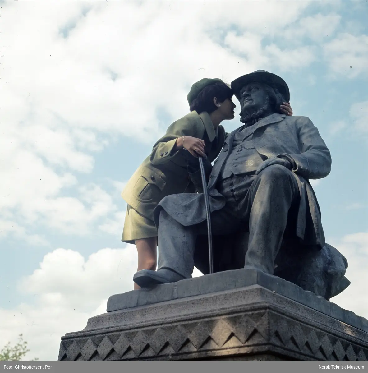 Motebilde, kvinne i grønn drakt, sittende på statuen av eventyrfortelleren Peter Chr. Asbjørnsen (1891) av Brynjulf Bergslien på St. Hanshaugen i Oslo, 1968. Bildet er en del av en serie der noen er publisert i ukebladet Det Nye samme høst med billedteksten: "Øverst til venstre ser du en grønn melert drakt av 70% ull og 30% nylon/rayon. Modelen har svakt skrådd skjørt og lang jakke med belte, utenpålommer med runde hjørner og sesongens tradisjonelle skilpaddespenne og knapper. Modellen koster ca kr. 400,-"