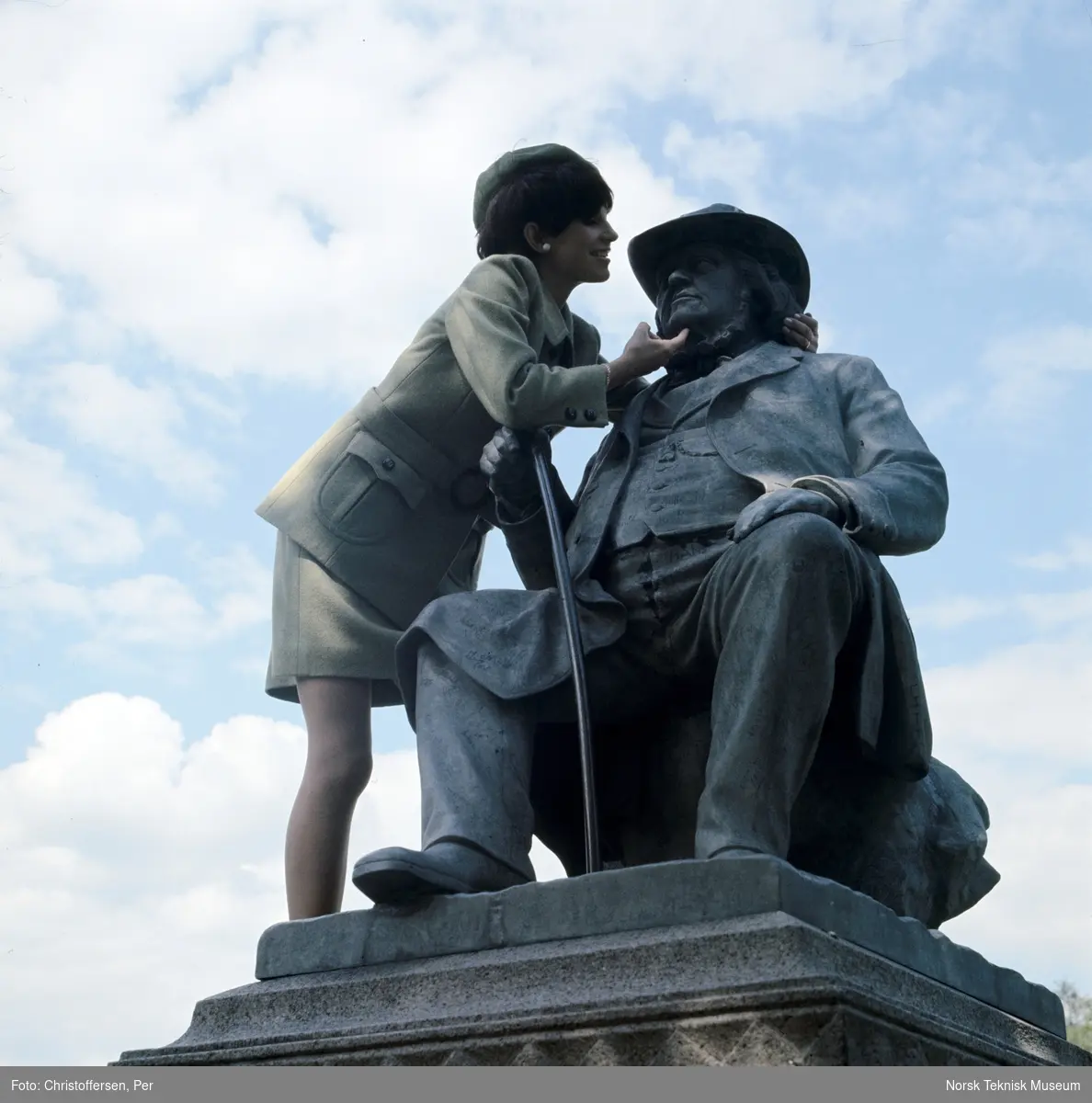 Motebilde, kvinne i grønn drakt, sittende på statuen av eventyrfortelleren Peter Chr. Asbjørnsen (1891) av Brynjulf Bergslien på St. Hanshaugen i Oslo, 1968. Bildet er en del av en serie der noen er publisert i ukebladet Det Nye samme høst med billedteksten: "Øverst til venstre ser du en grønn melert drakt av 70% ull og 30% nylon/rayon. Modelen har svakt skrådd skjørt og lang jakke med belte, utenpålommer med runde hjørner og sesongens tradisjonelle skilpaddespenne og knapper. Modellen koster ca kr. 400,-"