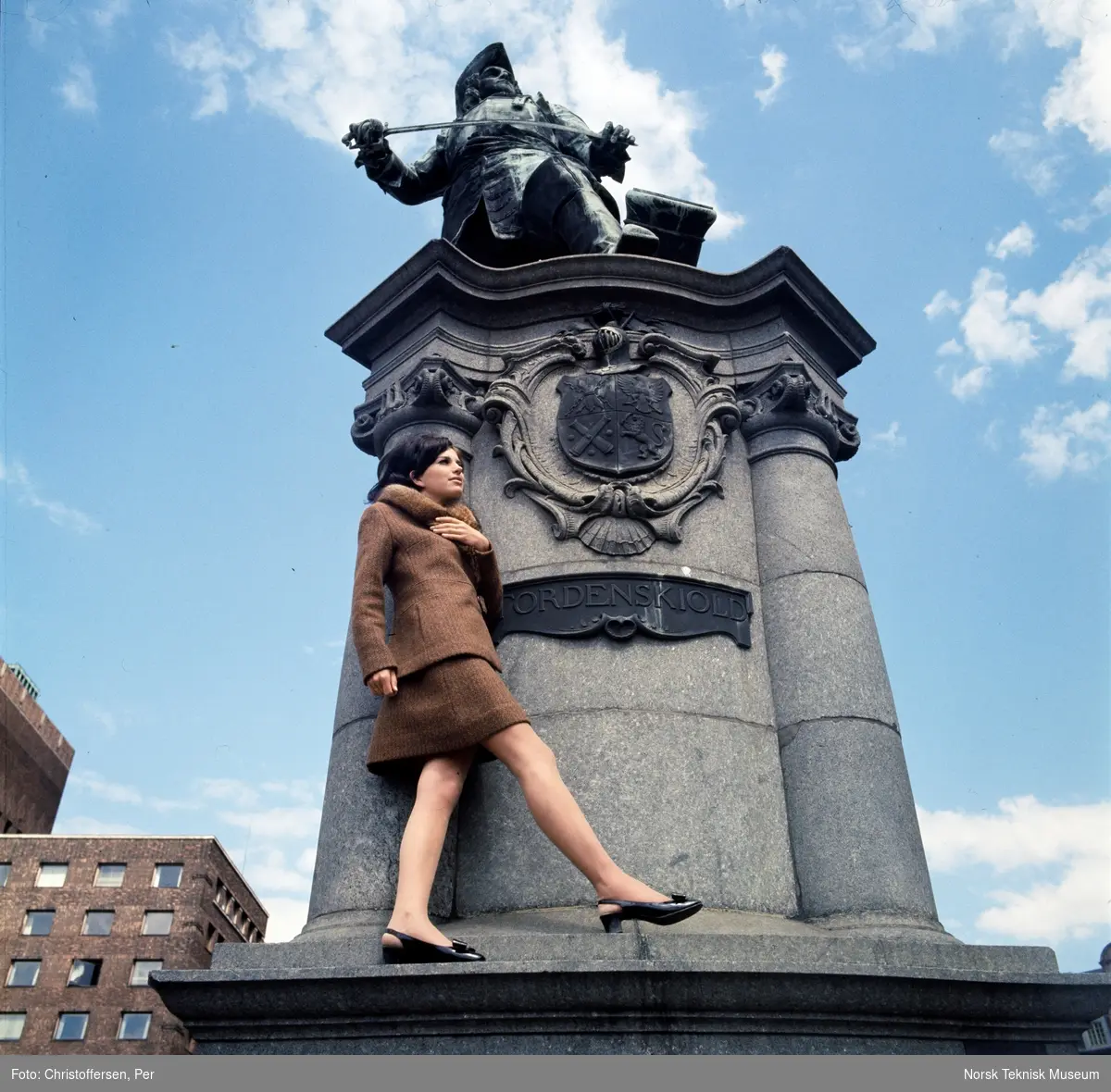 Motebilde i farger. Kvinnelig modell i todelt brun ulldrakt med pelskrage, foran statuen av Peter Wessel Tordenskiold (1901) av Aksel Ender på Rolf Strangers plass/ Rådhusplassen i Oslo, 1968. Bildet er en del av en serie der noen er publisert i en motereportasje i ukebladet Det Nye samme høst.