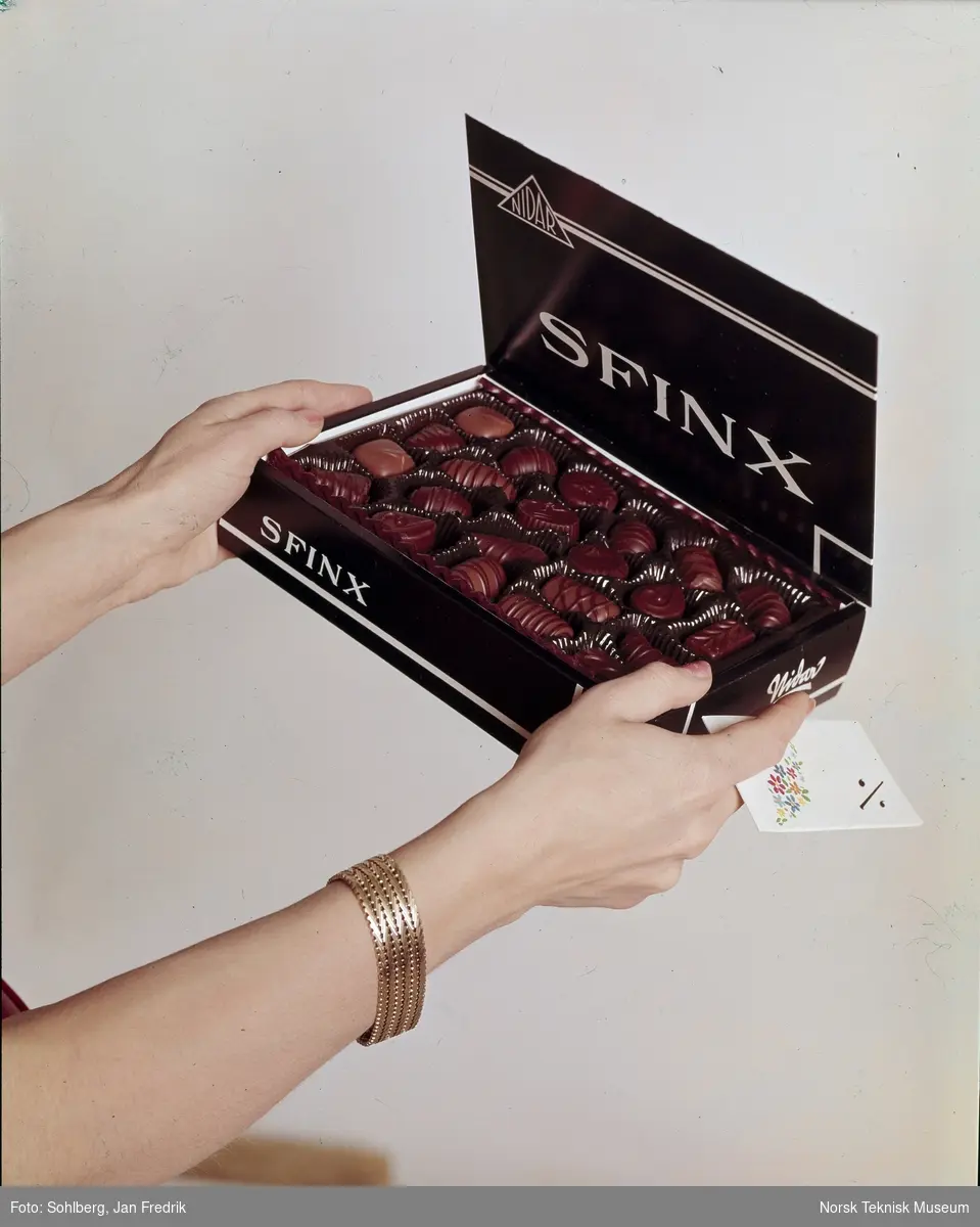 Reklamefotografi for konfektsjokolade. To kvinnehender holder fram en eske med Sfinx-konfekt og et kort.