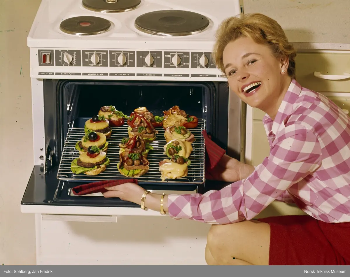 Reklamefoto for komfyr. Med moderne kjøkkenutstyr er livet som husmor ganske enkelt. En smilende kvinnelig modell lager lune smørbrød i stekeovnen.