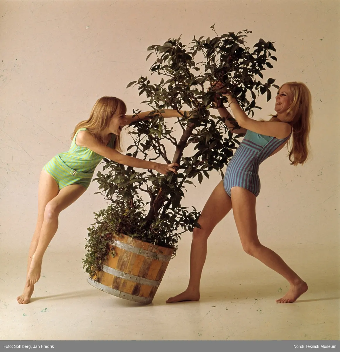 To kvinnelige modeller viser bademote, begge har badedrakt. De er fotografert ved en busk, plantet i en tønne. Bildet er del av en serie trykt i Det Nye. Bildet er merket  DN 25 s. 26-27 / 5 / K