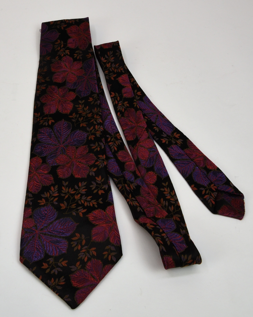 Mönstrad slips med svart botten. Mönstret består av broderade blommor och lövverk i bland annat violett och rosa, mönstret upprepas längs hela slipsen. Slipsen är fodrad med en röd textil med grå text.