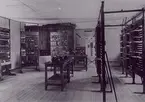 Nya AB Autotelefon Betulander Stockholm (Liljeholmen) april 1915. Provanläggning för mellanledningssystem (reläväxel), uppbyggd som förslag till automatisering av Stockholms telefonnät. De två stativraderna till höger representerar en huvudstation och de separata stativen till vänster tre understationer. Anläggningen demonstrerad för Telegrafstyrelsen och Stockholmstidningarna. (Pressvisningen ägde rum 26.4.1915). Beskrivning i bland annat Ericssons Review 1947 sid. 68-74.