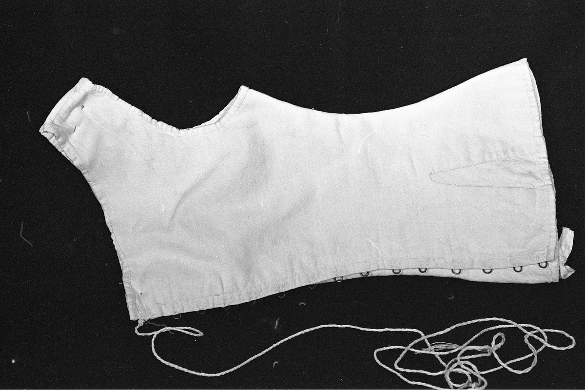 Liv eller korsett av lin med metallhekter og tvunnet tråd til snøring. Det har søm midt bak med hempe oppe og nede. Sidesømmen er litt tilbaketrukket, og det er felt inn stoff neders i sida for utsving fra livet. Halsåpningen er båtformet.