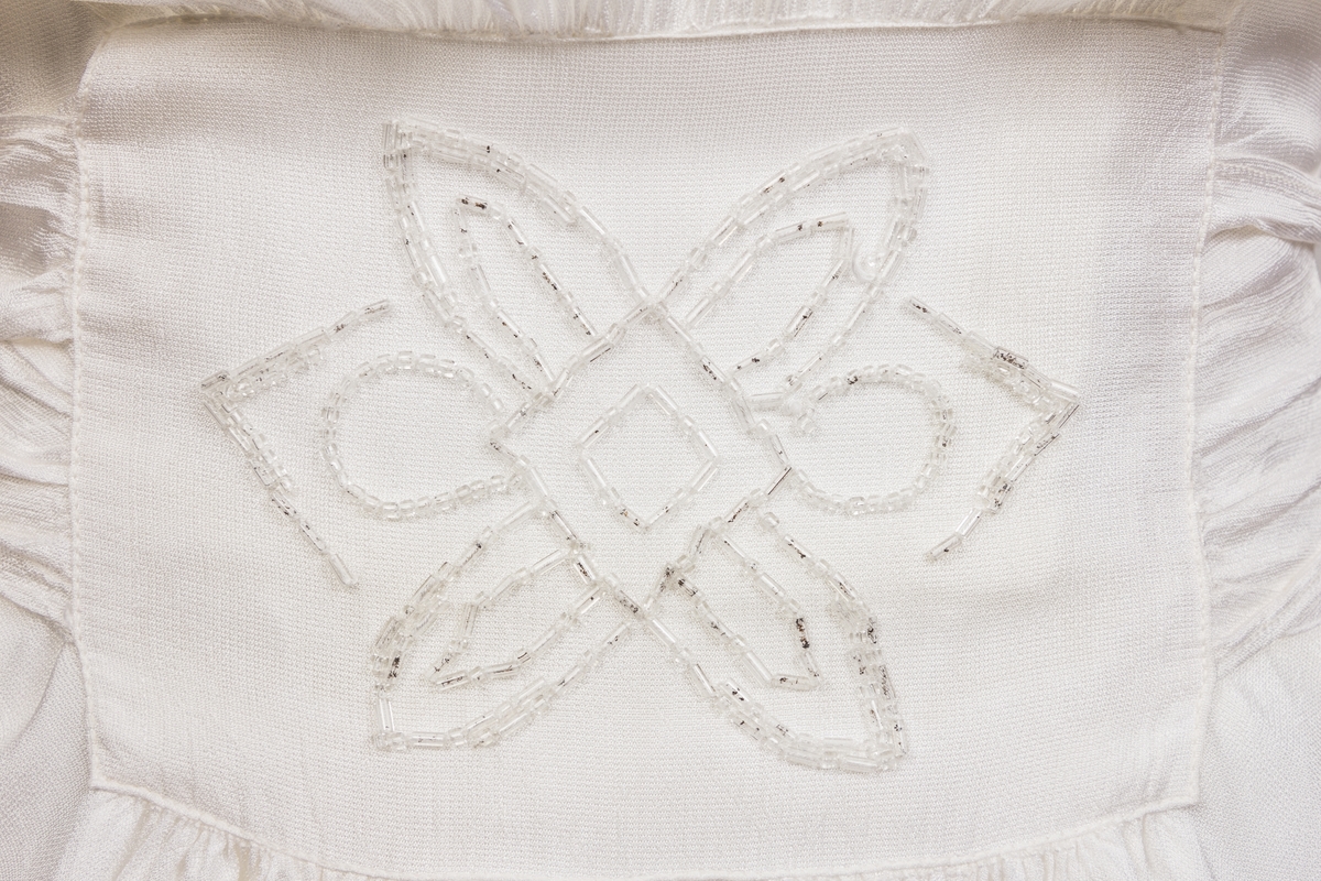 Konfirmasjonskjole, silkekjole dekorert med perlebroderier i front i midje, på skulderpartier og mansjetter. Kjolen er sid med kort erm og lukkes med glidelås og trykkapp i venstre side. Påsydd bredt bånd fra perlebrodert stykke i front som er festet bak med fire knapper.