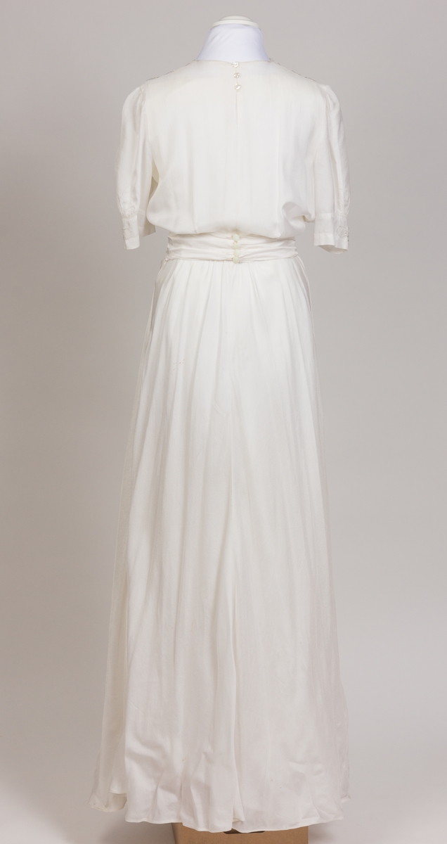 Konfirmasjonskjole, silkekjole dekorert med perlebroderier i front i midje, på skulderpartier og mansjetter. Kjolen er sid med kort erm og lukkes med glidelås og trykkapp i venstre side. Påsydd bredt bånd fra perlebrodert stykke i front som er festet bak med fire knapper.