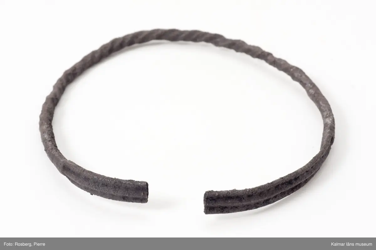 KLM 4975. Armring, av silver. Helt svartkorroderad, möjligen skadad av eld. Har varit sluten men är nu bruten. Något över hälften består av en rund, snodd ten, resten av en platt ten med tre längsgående, låga åsar. Två bitar av en annan dylik ring, bitarna består av den platta delen, hittades på samma ställe, vilka ingår under samma nummer.