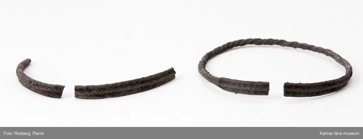 KLM 4975. Armring, av silver. Helt svartkorroderad, möjligen skadad av eld. Har varit sluten men är nu bruten. Något över hälften består av en rund, snodd ten, resten av en platt ten med tre längsgående, låga åsar. Två bitar av en annan dylik ring, bitarna består av den platta delen, hittades på samma ställe, vilka ingår under samma nummer.