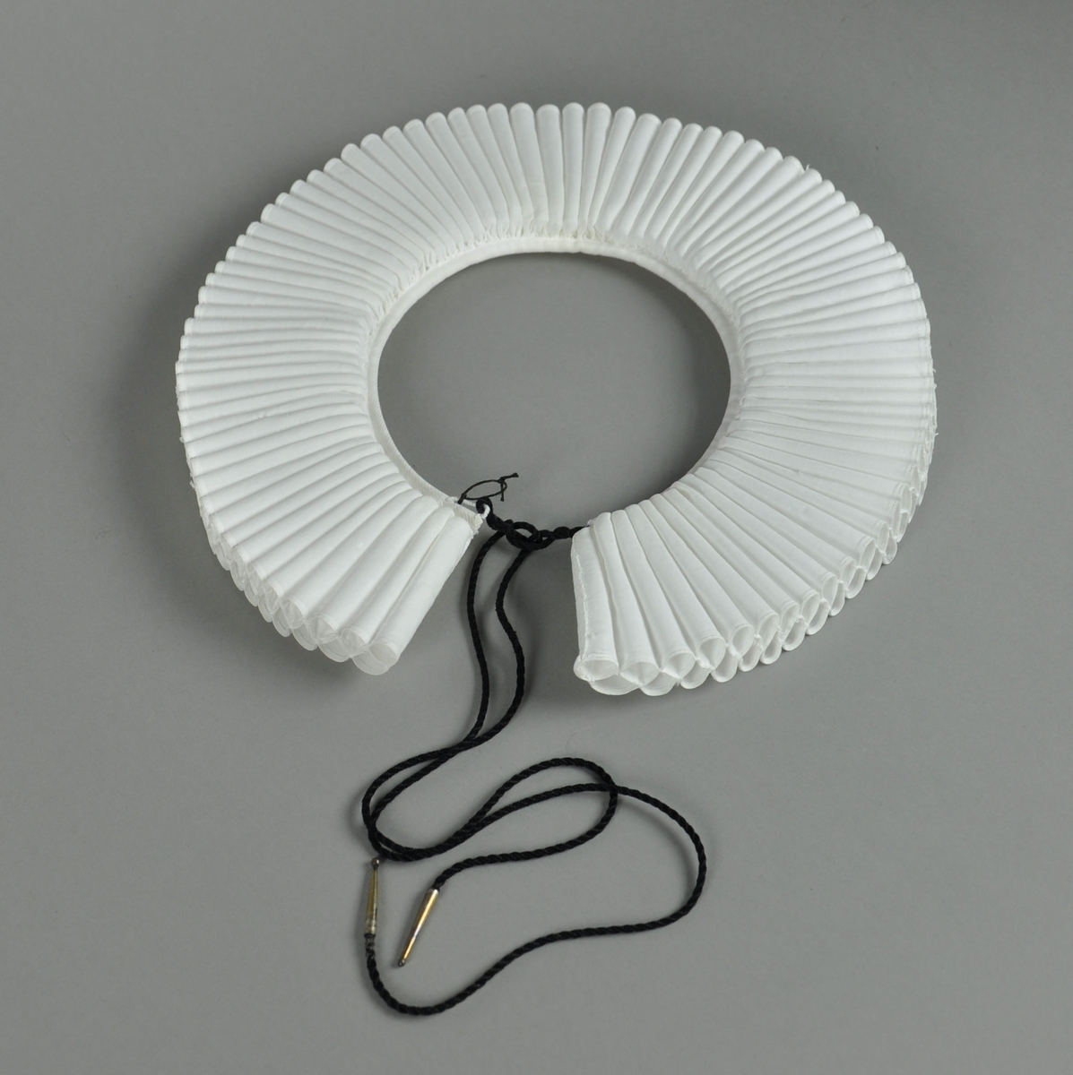 Hvit prestekrage av bomull. Kragen har 2 x 89 piper, 6,5 cm lange. Pipene et sammenfoldet bendelbånd med to hemper og svart messingtupper. Sammen med kragen hører en snipp, også hvit.