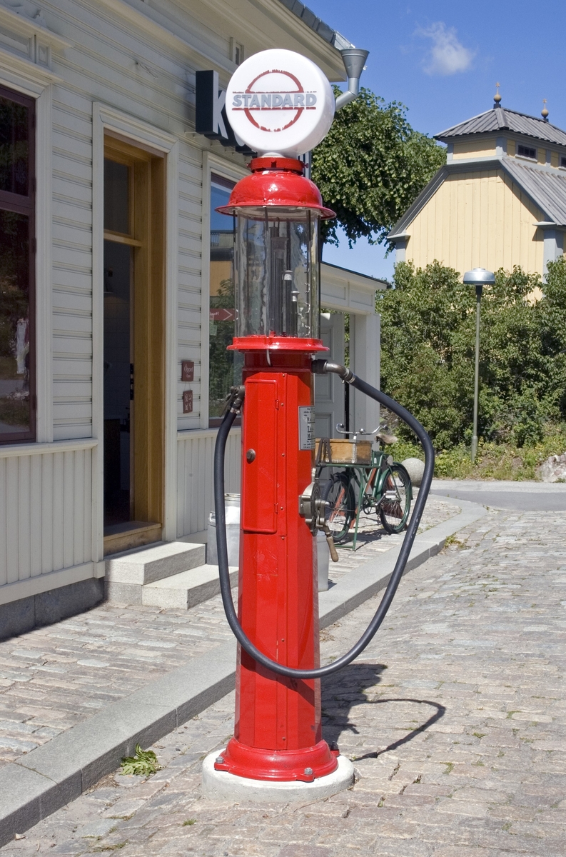 Bensinpump, fabriksgjord. Överst sitter en glasglob, SKAFOR.0015125 på ett metallfäste.  Under globen en cylinder av glas med mätmekanism och gradering. Därunder kommer det rödlackade pumphuset av stål. På en sida sitter en etikett av metall med text: Enligt Kungl. Maj:ts förordning angående eldfarliga oljor är det Förbjudet [understruket] att härstädes Röka Tobak [understruket] samt Tända [understruket] eller Medföra Eld [understruket]. Den som bryter mot sagda föreskrift, äventyrar jämlikt förordningen angående eldfarliga oljor § 60 ansvar av böter från och med 10 t.o.m. 500 kr." Etiketten sitter strax ovanför ett pumphandtag med omgivande dekorativt utformade "bricka" av metall och handtag svarvat av trä. "Brickan" är rund och utformad med graderingar från 0 till 25 med femliters intervaller. Text: PUMP IV-SC 54 A.B. J.C. LJUNGMAN MALMÖ VICI No 6705, ett räkneverk och LITER. Till höger om detta sitter ett annat pumphandtag av rödlackerat stål, fäst nertill. Ovanför etiketten börjar bensinslangen av gummi i ett metallfäste. Den fortsätter i en böj neråt till vänster, runt och upp och avslutas av den utgående kranen av metall, som hänger i ett hål på cylindern. Där bredvid finns också en låsbar lucka.