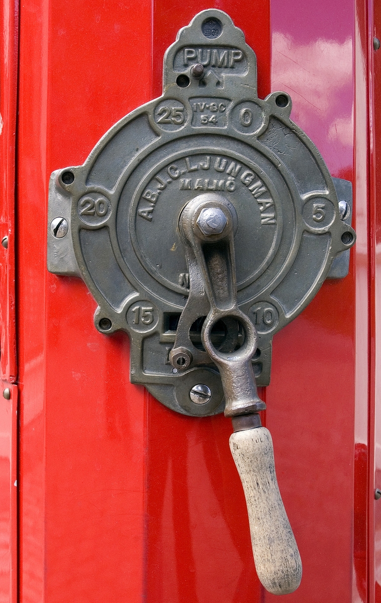 Bensinpump, fabriksgjord. Överst sitter en glasglob, SKAFOR.0015125 på ett metallfäste.  Under globen en cylinder av glas med mätmekanism och gradering. Därunder kommer det rödlackade pumphuset av stål. På en sida sitter en etikett av metall med text: Enligt Kungl. Maj:ts förordning angående eldfarliga oljor är det Förbjudet [understruket] att härstädes Röka Tobak [understruket] samt Tända [understruket] eller Medföra Eld [understruket]. Den som bryter mot sagda föreskrift, äventyrar jämlikt förordningen angående eldfarliga oljor § 60 ansvar av böter från och med 10 t.o.m. 500 kr." Etiketten sitter strax ovanför ett pumphandtag med omgivande dekorativt utformade "bricka" av metall och handtag svarvat av trä. "Brickan" är rund och utformad med graderingar från 0 till 25 med femliters intervaller. Text: PUMP IV-SC 54 A.B. J.C. LJUNGMAN MALMÖ VICI No 6705, ett räkneverk och LITER. Till höger om detta sitter ett annat pumphandtag av rödlackerat stål, fäst nertill. Ovanför etiketten börjar bensinslangen av gummi i ett metallfäste. Den fortsätter i en böj neråt till vänster, runt och upp och avslutas av den utgående kranen av metall, som hänger i ett hål på cylindern. Där bredvid finns också en låsbar lucka.