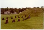 Badelunda sn, Anundshögsområdet, Långby.
Skeppssättning och gravhög med byggnader i bakgrunden.