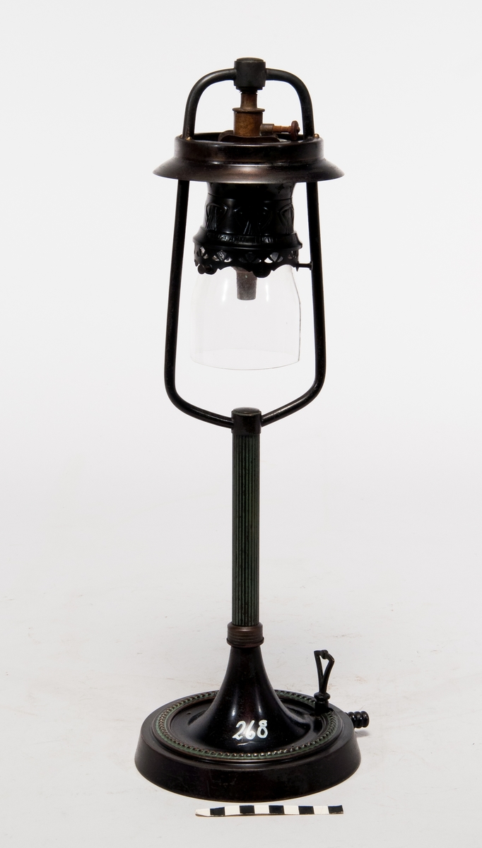 Gaslampa av bordsmodell i metall och glas, märkt: "Graftzin-light D.R.P. 126135". Lös ettikett med text: "Bordstake på svart fot. Bordstakarna härleda sig från 1870-1880 talet, var i bruk intill 1910 talet".
Påmålad märkning "268".