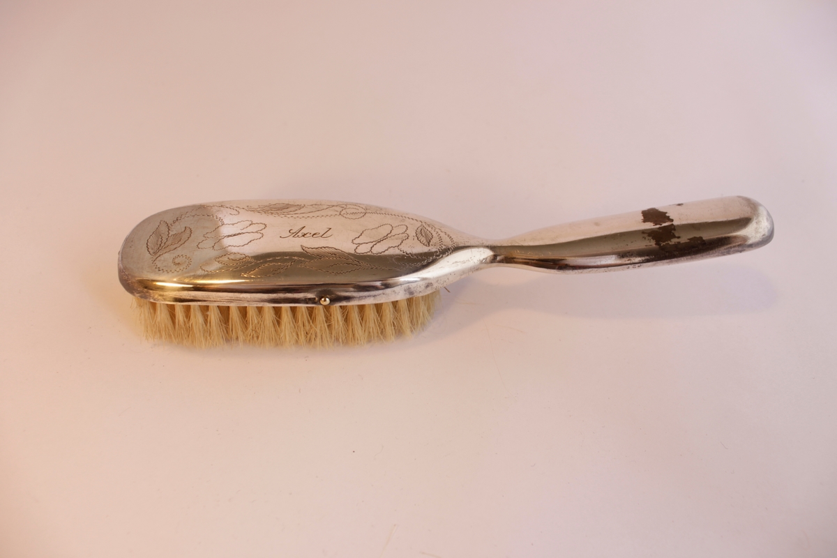 Borstgarnityr bestående av tre borstar en (a) hårborste, två klädesborstar (b-c). Naturborst. Borstarna är klädda i nysilver som har ingraverade stiliserade blommönster. Hårborsten och den ena klädborsten har namnet "Axel" ingraverat på ovansidan. Själva borsthuvudena är tillverkade av trä och sedan har en plåt av nysilver spikats på.