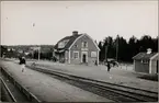 Vy på stationsområde och stationshus med resande på perrong.