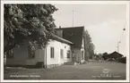 Vy på stationshus med parkering och lagerbyggnad i bakgrunden.