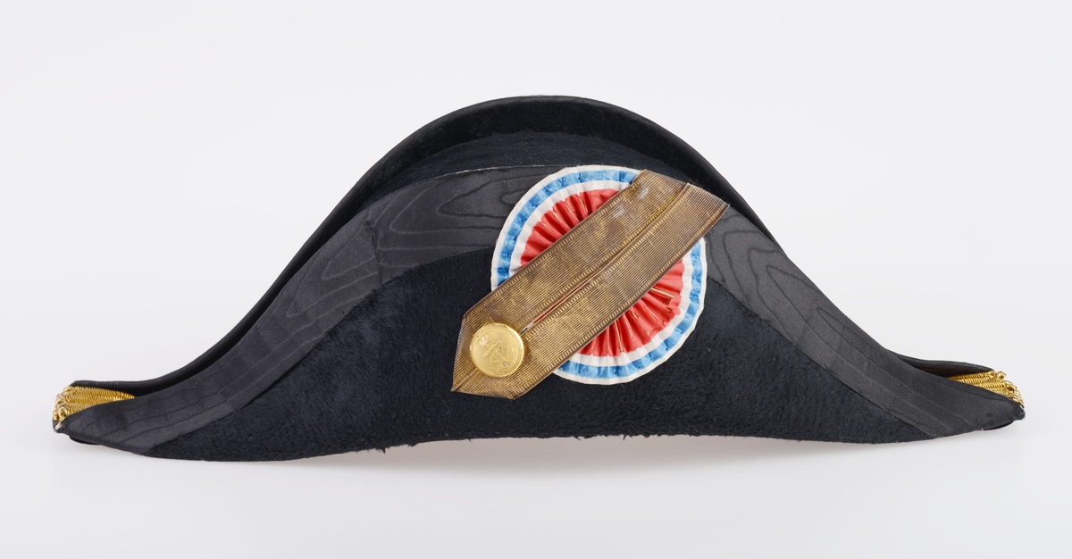 En hatteboks (FTT.56303.01) som inneholder en snutehatt (FTT.56303.02), et bandolær (FTT.56303.03) og en epålett (FTT.56303.04).  
FTT.56303.01:
Hatteboksen er laget av blikk og er svartlakkert utenpå. Lokket er hengslet på baksiden. Oppå lokket er det et enkelt håndtak. I fronetn er det en hempe for hengelås. Den har to lave bein som er laget av sammenrullet blikk. Boksen har form tilpasset hatten. Esken har ikke fôr, men det ligger et papir i bunnen.
FTT.56303.02:
En svart uniformshatt, nærmere bestemt en snutehatt eller en tosnutet hatt. Det er gullbånddekor på hver snute. På den ene siden er det festet en stor kokarde i rødt, hvitt og blått. Over denne er det to flate gullbånd som ender i en spiss. I enden er det en gullfarget metallknapp med et anker med en kongekrone over. Hatten er laget av svart silkefloss med svarte silkebånd langs kanten. Hatten har hvitt silkefôr og svettebånd av brunt lær. På svettebåndet er det festet brune skinnreimer til å feste under haken. På den ene siden er det festet en hvit silkesløyfe. Silkefôret har informasjon om produsent, se "Påført tekst/merker". Det er også bilde av en edderkoppe og et edderkoppnett. Hatten har avstivning ved hjelp av papp. Hatten er håndsydd.
FTT.56303.03:
Et bandolær laget av svart skinn, svart silkestoff og messing. Midjebeltet har en spenne midt foran. Spennen har motiv av et anker med en krone over. På innsiden av beltet er det sydd fast en skinnreim med metallhull til å regulere livvidden. Skulderreimen er festet i midjebeltet med metallringer. Delene til skulderreimen festes sammen med to fjærbelastende kroker. Ved sammenføyningen skulderreim og midjebelte er det påmontert en liten kjetting med krok.
FTT.56303.04:
En skulderklaff til uniform. Oversiden består av svarte- og gullfargede flettede snorer. På undersiden er det sydd på svart ullstoff som er like bred som klaffen. I den ene enden er det sydd på en svart knapp som ullstoffet kan festes i.